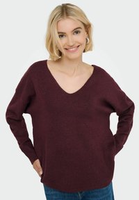 Burgundska pletena jopica z V-izrezom, dolgimi rokavi in rebrastimi manšeti. Mehka tekstura in sproščen kroj, v kombinaciji z modrimi kavbojkami.