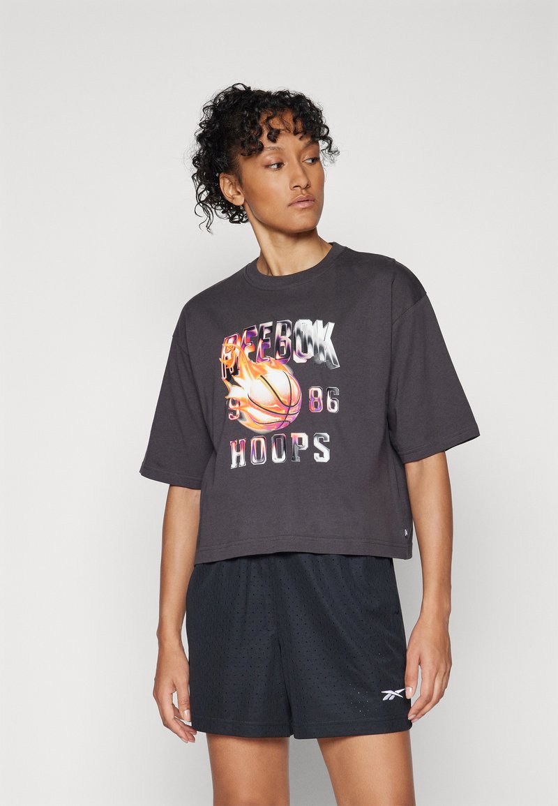 Camiseta corta de color gris oscuro con un gráfico de baloncesto y el texto "Reebok 96 Hoops"; combinada con pantalones cortos deportivos de malla negra que presentan perforaciones sutiles.