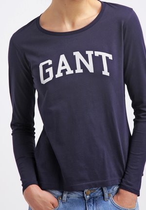 Marineblauw shirt met lange mouwen met wit "GANT"-logo, gedragen met lichtblauwe spijkerbroek, handen gedeeltelijk in de zakken, torso zichtbaar.