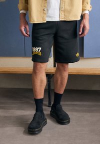 Black athletic shorts with yellow "1897 JUVENTUS" text, paired with black slip-on shoes and dark socks. The outfit features a casual design.