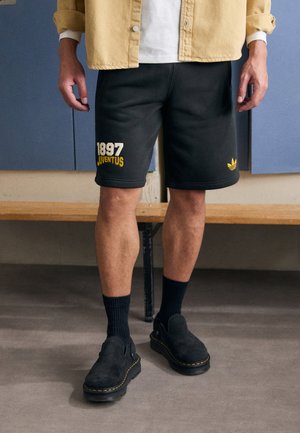 Črne športne hlače s rumenim napisom "1897 JUVENTUS", v kombinaciji s črnimi klompli in temnimi nogavicami. Obleka ima sproščen dizajn.