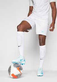 Light blue Nike football boots on a white leg, paired with white shin guards, shorts, and a shirt, standing on a black and white patterned ball.