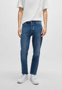 Dark blue denim jeans with a straight leg fit, five-pocket design, light stitching, and a button closure at the waist. Worn with white sneakers.