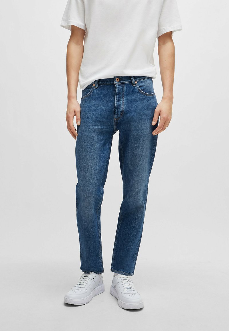 Dark blue denim jeans with a straight leg fit, five-pocket design, light stitching, and a button closure at the waist. Worn with white sneakers.