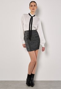 Blusa blanca de manga larga con cuello con volantes y corbata negra; falda corta ajustada de textura metálica gris; botas negras elegantes hasta el tobillo.