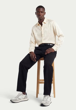 Uomo seduto su sgabello di legno che indossa una camicia crema a maniche lunghe con texture, pantaloni neri, cintura nera e scarpe da ginnastica bianche su sfondo chiaro.