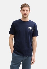 Námořnická modrá bavlněná tričko s krátkými rukávy a kulatým výstřihem. Má potisk na hrudi s texty "BLUE" a "TOM TAILOR" v bílé barvě.