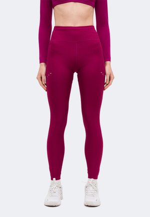 Un modèle porte un legging texturé taille haute de couleur magenta et un crop top assorti, avec des baskets blanches, devant un fond uni et clair.