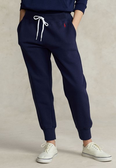 Personne portant un pantalon de jogging bleu marine avec un cordon blanc et un logo rouge, associé à des baskets blanches, debout sur un fond neutre.