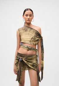 Frau in einem asymmetrischen Crop-Top in Beige und Braun sowie einem passenden Wickelrock, der gebunden ist. Sie trägt große Creolen und hat ihr Haar zurückgebunden.