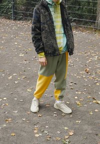 Fleecejacket in Dunkelblau mit schwarzem Muster, über einen hellgrünen Hoodie getragen. Hellgrüne und gelbe, colorblockierte Jogginghosen. Beigefarbene Sneakers.