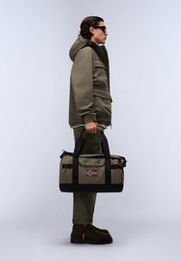 Sac de sport vert olive avec des accents noirs, des poignées et un écusson du drapeau norvégien. Matériau doux, forme cylindrique, avec des détails de fermeture éclair et de boucle.