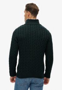 Pull en maille torsadée vert foncé avec un col montant et des poignets côtelés, porté par-dessus une chemise à carreaux, mettant en valeur des motifs torsadés texturés.