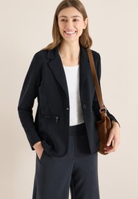 Zwart, op maat gemaakte blazer met een enkele knoop, ritssluitingen op de zakken en een gladde textuur, gecombineerd met een wit shirt en donkere broek. Bruine handtas.