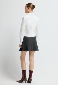 Vrouw staat met haar rug naar de camera, gekleed in een witte blouse met lange mouwen, een geplooide donkergrijze mini-jurk, bordeauxrode doorschijnende sokken en metallic puntige hakken.