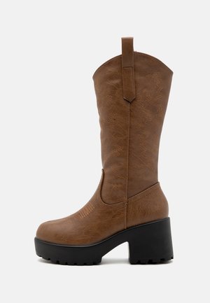 Botte haute en similicuir marron jusqu'au genou avec un bout pointu, des coutures contrastées et un talon noir épais. Présente des motifs décoratifs sur la tige.