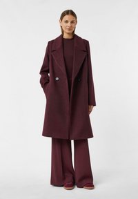 Bordeauxfarbener Wollmantel mit doppelreihigem Design, breiten Revers und seitlichen Taschen, kombiniert mit passenden weit geschnittenen Hosen und Schuhen.