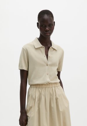 Donna dalla pelle scura che indossa una camicia beige a maniche corte con bottoni e una gonna beige a vita alta con fascia elastica, in piedi davanti a uno sfondo bianco.
