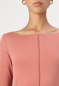 Haut à manches longues rose à côtes avec un décolleté asymétrique et des détails de couture centrale. Tissu lisse avec une texture douce.