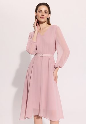 Kobieta w jasnoróżowej sukience midi z długim rękawem, dekoltem w serek i dopasowanym paskiem, pozująca na jednolitym jasnym tle.