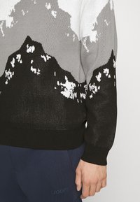 Maglione lavorato a maglia con motivo montano in nero, grigio e bianco. Maniche e orlo a coste, con una vestibilità ampia. Indossato con pantaloni blu scuro.