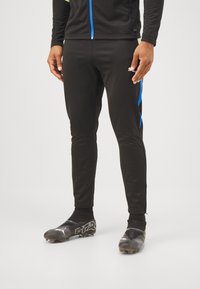 Pantalones deportivos negros con rayas laterales azules, hechos de una tela elástica. Combinados con botas de fútbol negras que presentan un diseño distintivo.