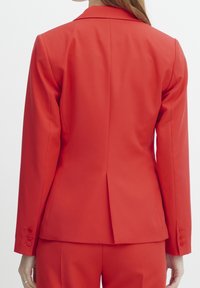 Blazer rouge cintré avec un revers cranté, une silhouette ajustée et une fente unique à l'arrière. Détails de boutons sur les poignets.
