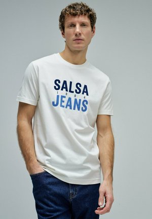 Hombre con pelo rizado con una camiseta blanca "Salsa Jeans 1994" y vaqueros azules sobre un fondo gris claro liso.