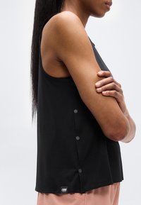 Haut sans manches noir avec des boutons décoratifs sur le côté, fabriqué dans un matériau lisse. La coupe est ample avec un ourlet arrondi.