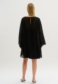 Schwarzes Kleid mit einem strukturierten Stoff und einem durchsichtigen Overlay, weiten Ärmeln und einer Schlüssellochöffnung hinten. Sichtbarer Silberschmuck.