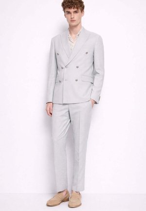 Giovane uomo con capelli ricci che indossa un abito doppiopetto grigio chiaro, una camicia floreale e mocassini beige, in piedi davanti a uno sfondo bianco.