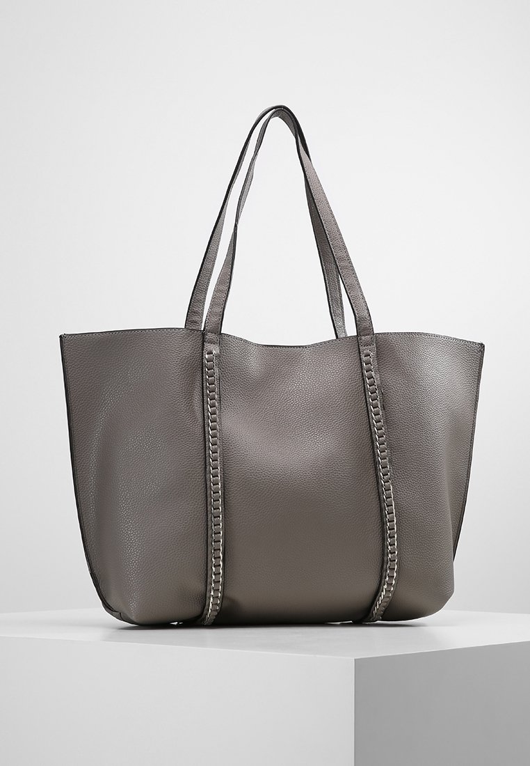 Sac fourre-tout en cuir gris texturé avec deux longues poignées et des détails de surpiqûres verticales, posé sur un piédestal blanc.