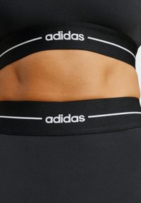 Black athletic top featuring a wide elastic band with white "adidas" branding and accent lines, smooth texture, designed for active wear.