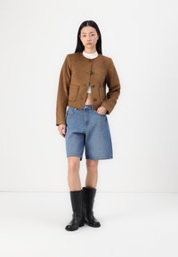 Brun cropped jakke med rund halsudskæring og store knapper, parret med højtaljede blå denimshorts og sorte knæhøje støvler.