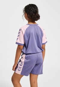 Tvådelad sportoutfit i nyanser av lila och rosa, med lodräta ränder och chevrondetaljer. Tillverkad av lätt, slät tyg.