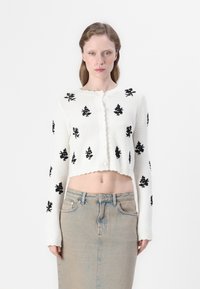 Cardigan bianco corto con ricami floreali neri, materiale a maglia testurizzato e bordi frangiati. Indossato sopra una gonna di jeans a vita alta.