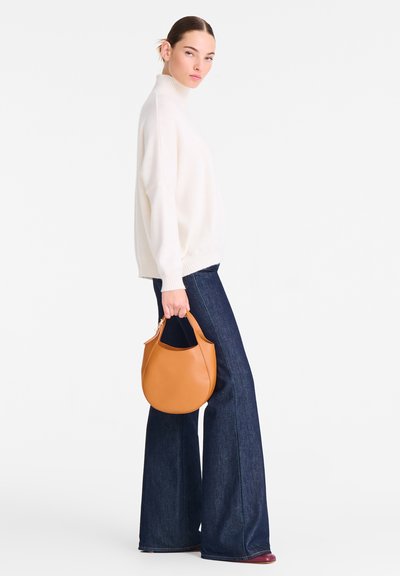 Hellbeiger Rollkragenpullover, locker geschnitten; dunkelblaue ausgestellte Jeans; kleine runde Handtasche aus cognacfarbenem Leder mit Henkel, glatte Oberfläche.