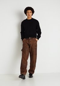 Μαύρο πουλόβερ, χαλαρού στυλ καφέ cargo παντελόνια με πλευρικές τσέπες, λεία υφή, κουμπωτή ζώνη και μαύρα παπούτσια. Ουδέτερο φόντο.