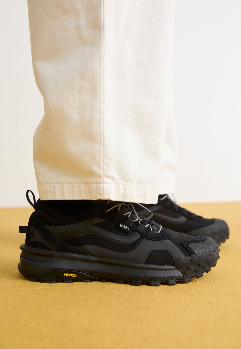 Zwarte sportieve schoenen met een gestructureerd bovenwerk, grijze veters en een Vibram-zool. G gedragen met lichtgekleurde, comfortabele broek.