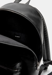 Zaino sintetico nero con un esterno liscio, apertura con cerniera e interni semplici. Presenta spallacci regolabili e un'etichetta con il logo all'interno.
