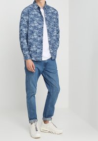 Blau gemustertes Hemd über einem weißen T-Shirt, kombiniert mit blauen Jeans und weißen Sneakers. Einfaches Design mit lässiger Passform.