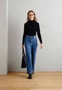 Svart polotröja, högmidjade blå jeans, svarta skor och en svart tote-väska. Trägolv och slät vägg i ljusgrå färg som bakgrund.
