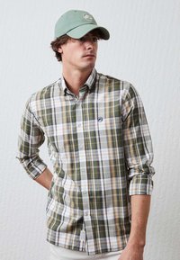 Camisa de cuadros verde, blanca y amarilla con botones, de manga larga, con los puños recogidos y un logo azul en el pecho. Tela ligera.
