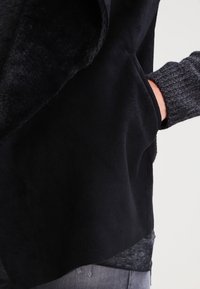 Osoba w ciemnej polarowej kurtce, z jedną ręką w bocznej kieszeni, nałożonej na szary sweter robiony na drutach i szare spodnie.