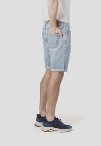 Hele sinised teksapüksid, millel on ülespööratud sääred ja kulunud detailid, kantud tumesiniste spordijalatsitega, millel on beež tall ja võrgust detailid.