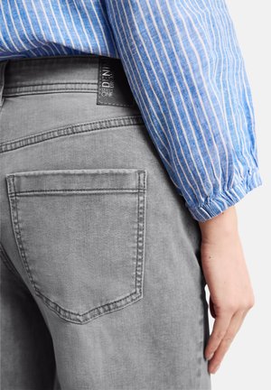 Persoon die een grijze spijkerbroek en een blauw-wit gestreept shirt met lange mouwen en elastische manchetten draagt, met zicht op het achterzakje en de hand aan de zijkant.