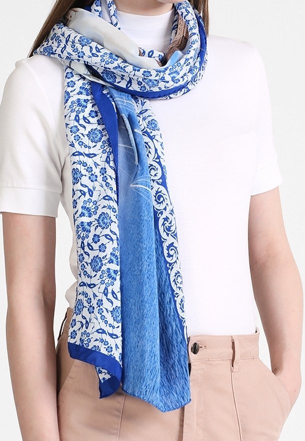 Femme portant un haut blanc à manches courtes et un pantalon beige, avec une écharpe bleue et blanche à motifs drapée autour du cou.