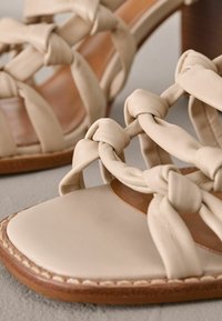 Des sandales à talons en cuir beige présentent des brides nouées, une semelle rembourrée et un talon en bois. La semelle a une texture tressée.