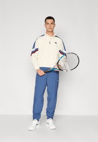 Balta zip-up striukė su mėlynais ir raudonais akcentais, derinama su mėlynomis treniruočių kelnėmis ir baltomis sportinėmis bateliais. Asmuo laiko teniso raketę.