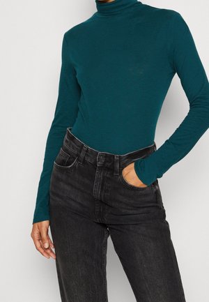 Personne portant un col roulé à manches longues couleur sarcelle, rentrée dans un jean noir taille haute, avec une main dans la poche.